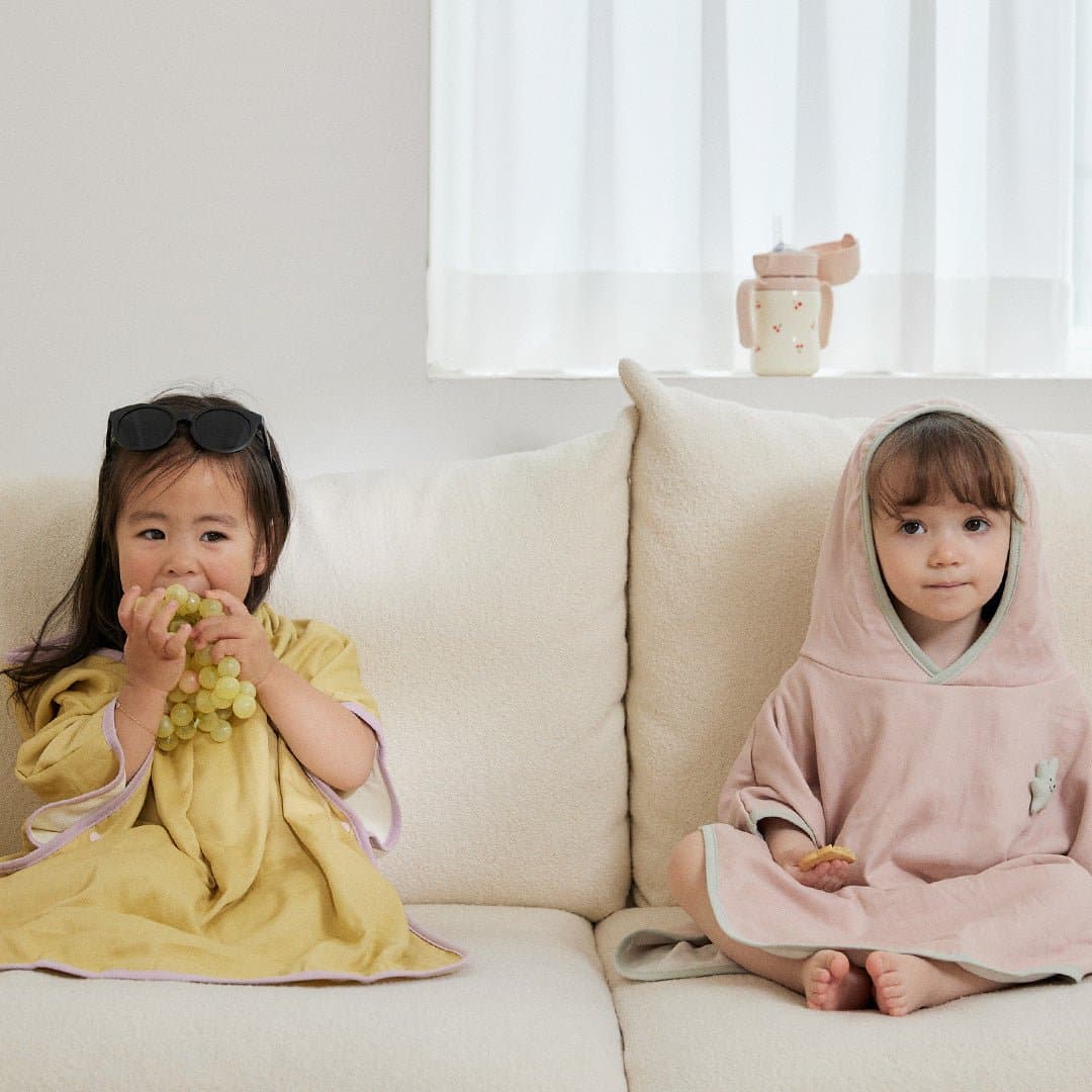 Two toddlers on a couch in bamboo poncho towels by a sunlit window
